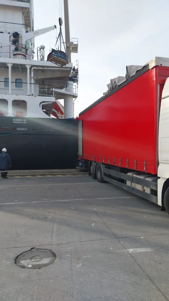 Camión rojo estacionado junto a un barco con grúa en operación.