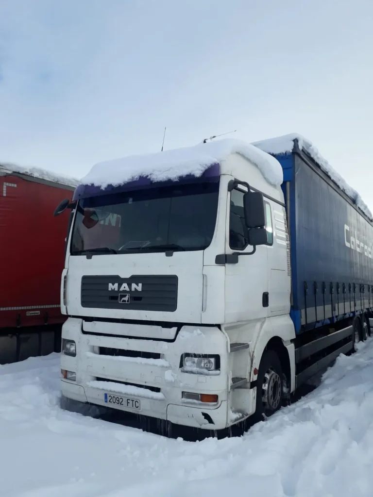 Camión MAN blanco cubierto de nieve estacionado junto a otro camión rojo.